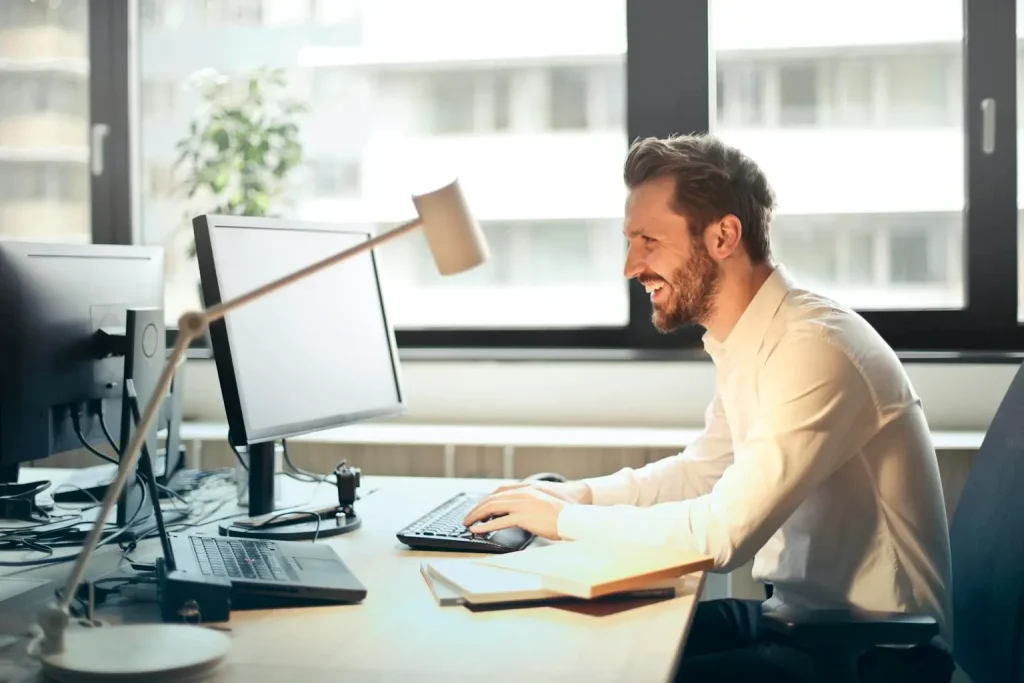 Confident Professional at Work in a Bright Modern Office Smiling man in a white shirt working at a desk with dual monitors in a bright, modern office.