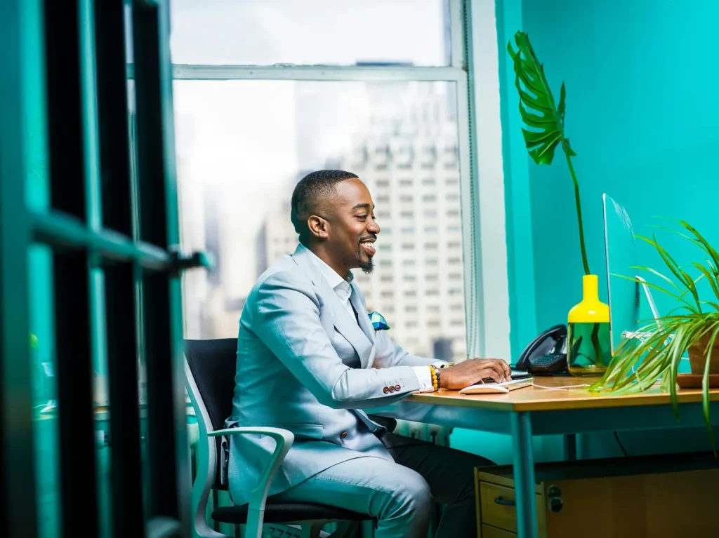 Confident Professional at Work in a Bright Modern Office Smiling businessman in a light blue suit working at a desk in a modern, brightly colored office with a city skyline in the background.