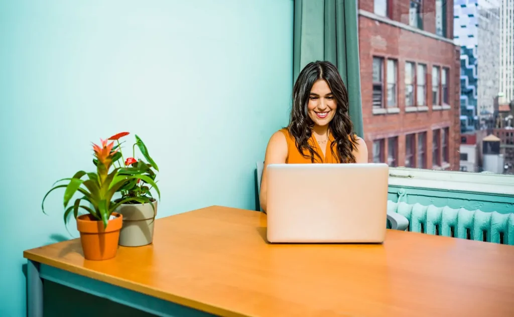 Productive Workday with a View in a Bright Urban Office Smiling woman in an orange top working on a laptop at a bright office desk with potted plants and a city view.