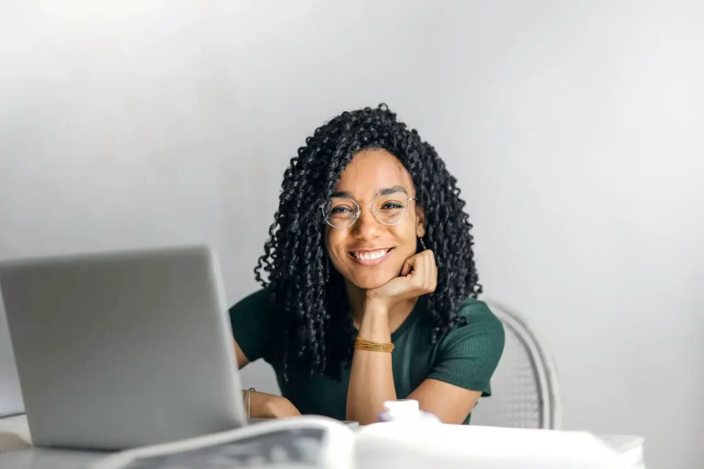 Confident woman working on a laptop at home or office Smiling woman with curly hair and glasses sitting at a desk with a laptop and open book.