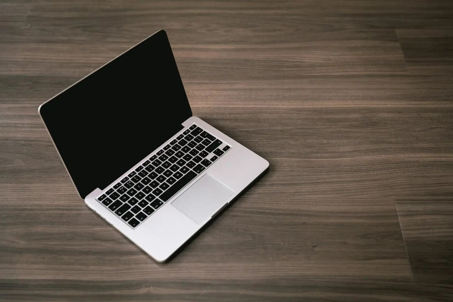 Businesswoman working on a laptop at a bright, organized desk with notebooks and a cup of coffee. Open silver laptop with a black screen placed on a smooth, dark wooden floor.