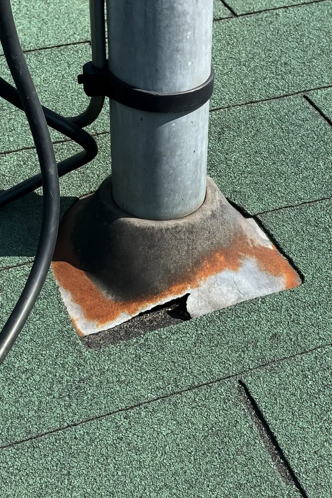 Close-up of a rusted roof flashing base around a metal pole on a green shingle roof during a repair by AIC Roofing & Construction.