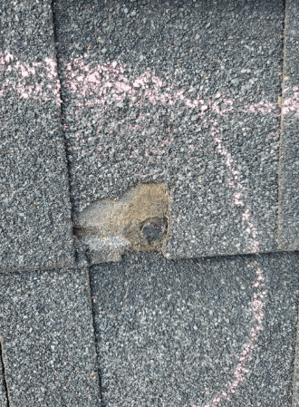 Damaged asphalt roof shingle with a puncture and chalk markings.