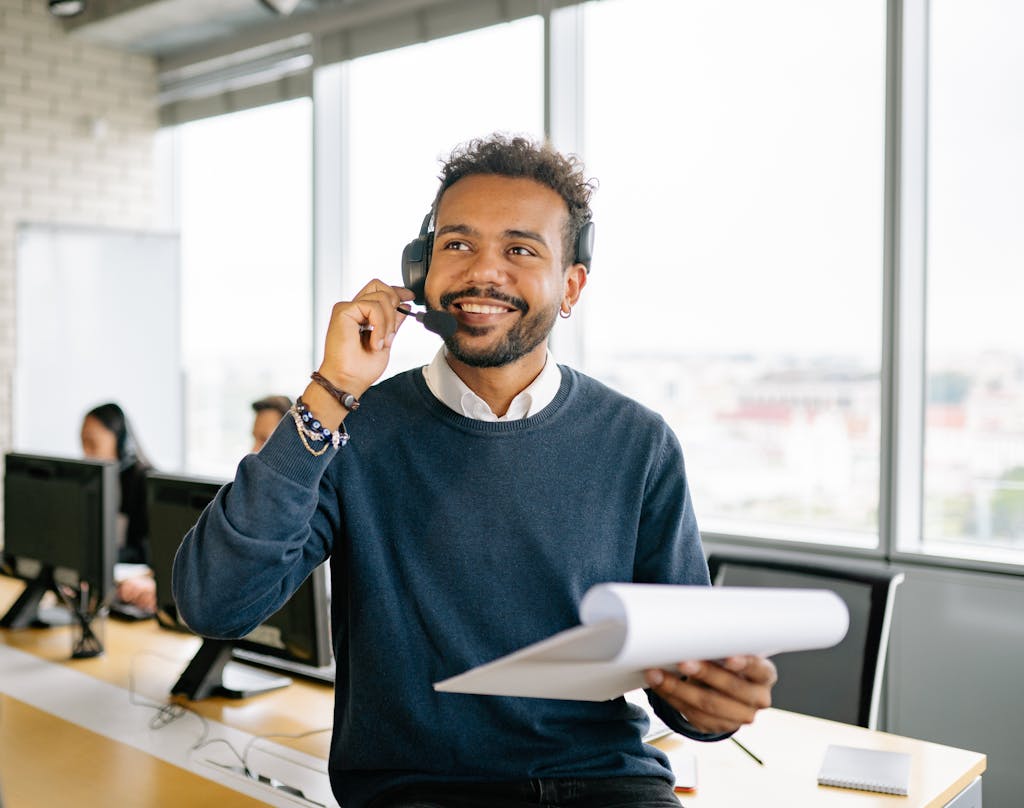 Confident Professional at Work in a Bright Modern Office A cheerful man wearing a headset and holding a clipboard at a modern office workspace.