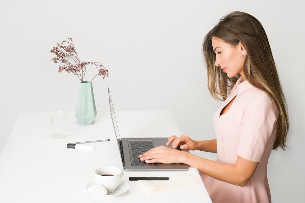 Confident Professional at Work in a Bright Modern Office A woman in a pink dress working on a laptop at a modern, minimalist desk.