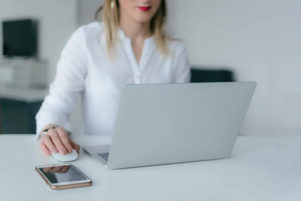 Professional Working Remotely at a Modern Desk Woman in a white blouse working on a laptop at a clean, modern desk with a smartphone beside her.