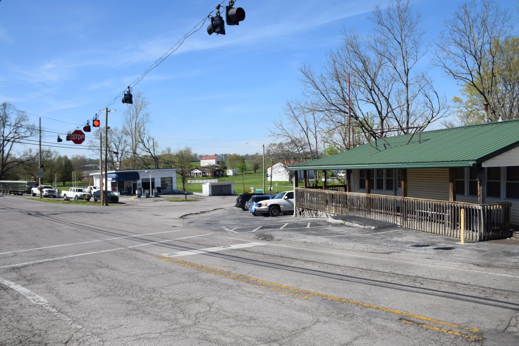 A rural street intersection in Burgin, KY with a green-roofed building in the foreground, a stoplight above, and a small gas station across the street.