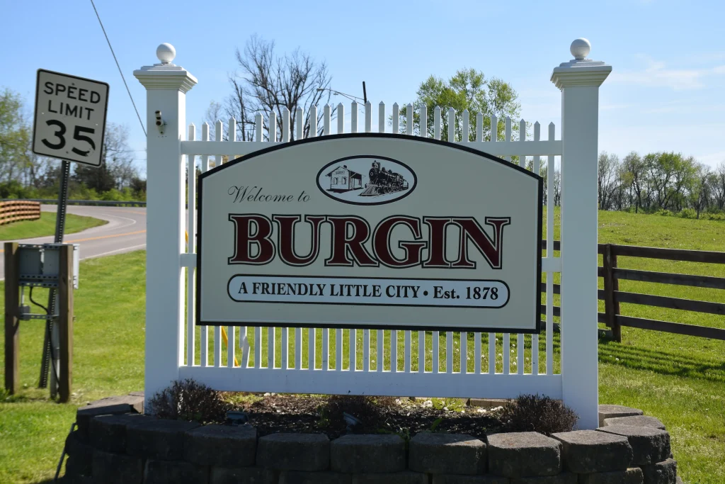 Welcome sign for Burgin, KY reading "A Friendly Little City • Est. 1878," placed in front of a white fence along a rural roadside.