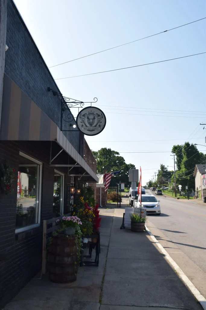Buffalo Spirits storefront with flowers and signage along a quiet street in downtown Stamping Ground, KY.