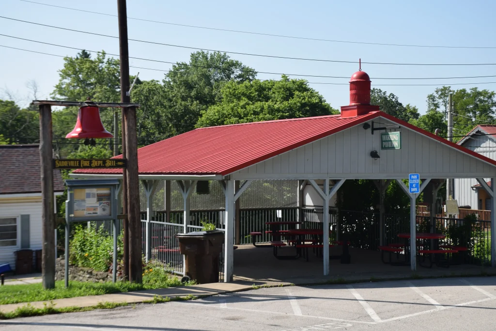 A covered public pavilion with a red metal roof in Sadieville, KY, featuring picnic tables and a historic fire department bell on display nearby.