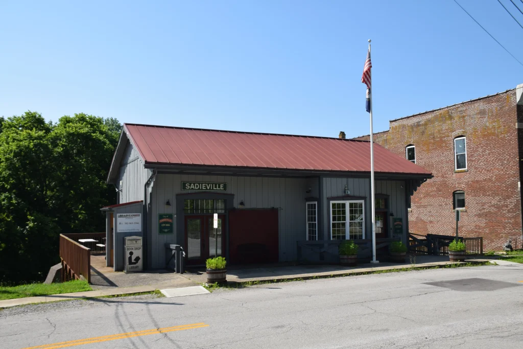 The Sadieville City Hall building with a red roof, gray siding, and American and Kentucky flags out front, located on a quiet street.