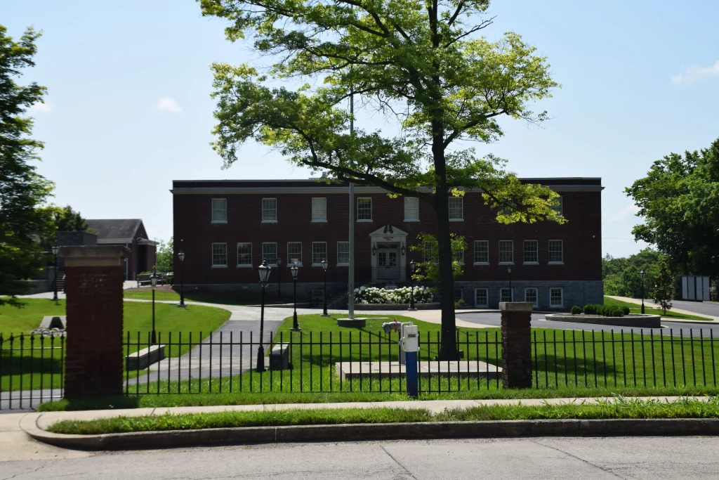 Brick school building with white trim and manicured lawn, viewed through a black iron fence on a sunny day.