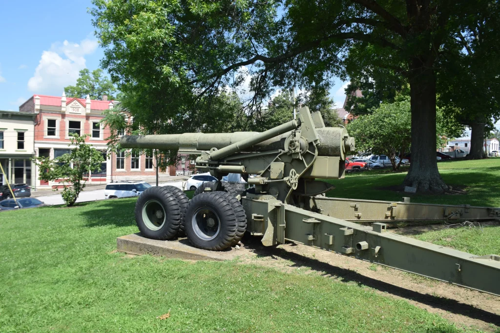 Military artillery cannon displayed on a lawn in Carlisle, KY with historic downtown buildings in the background.