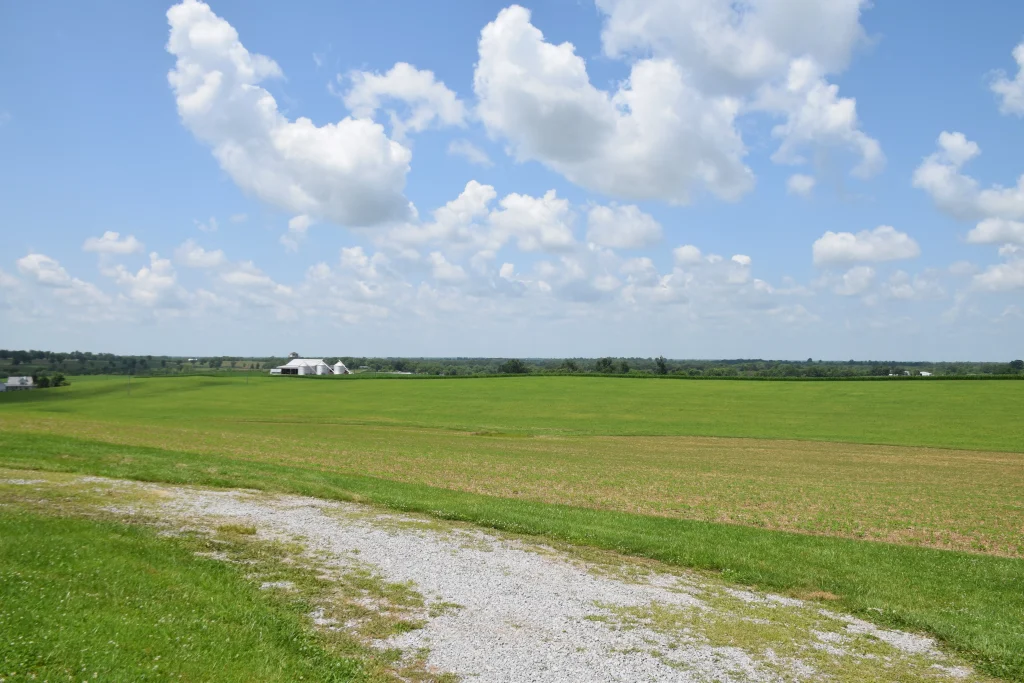 Open farmland in North Middletown, KY, with green fields, a gravel path, and a distant white barn under a blue sky with scattered clouds.