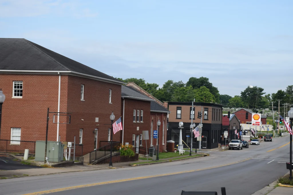 Main street in downtown Perryville, KY, lined with brick buildings, American flags, and small businesses, with a Shell gas station visible in the distance.