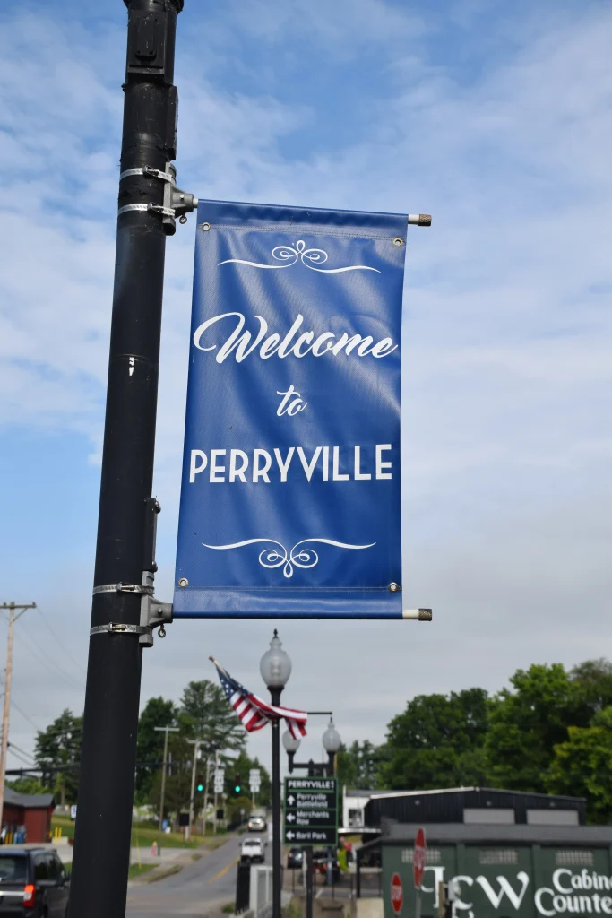 Blue street banner reading “Welcome to Perryville” attached to a black lamppost, with an American flag and downtown Perryville in the background.