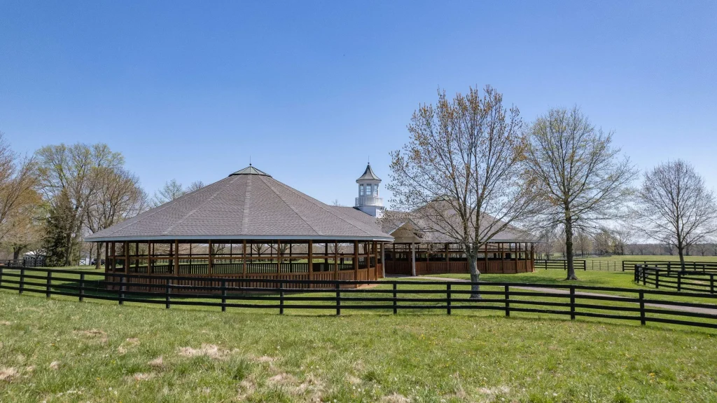 Large round training barn in Kentucky with a newly installed roof by AIC Roofing & Construction, surrounded by fencing, trees, and open pasture.