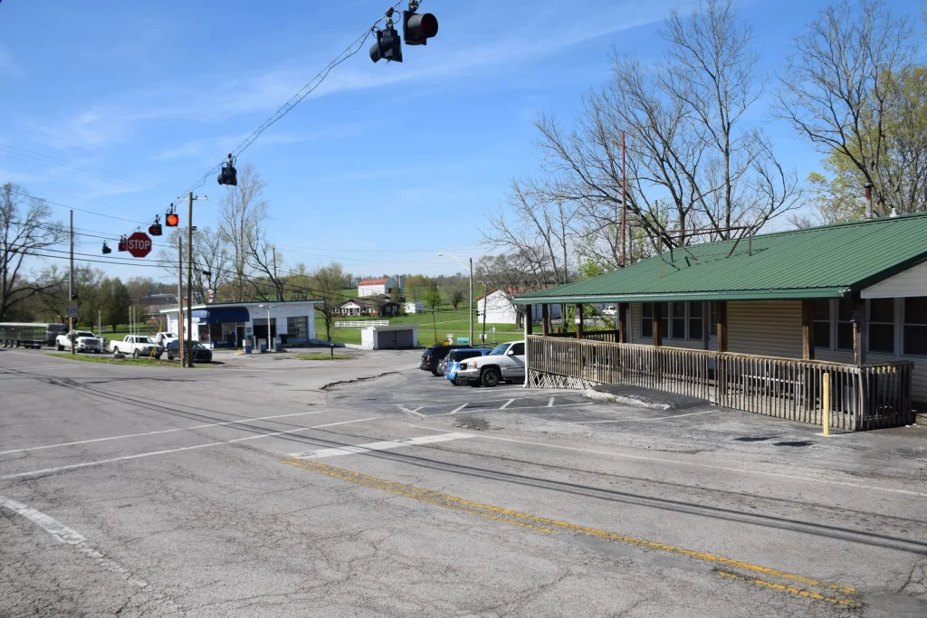 A rural street intersection in Burgin, KY with a green-roofed building in the foreground, a stoplight above, and a small gas station across the street.