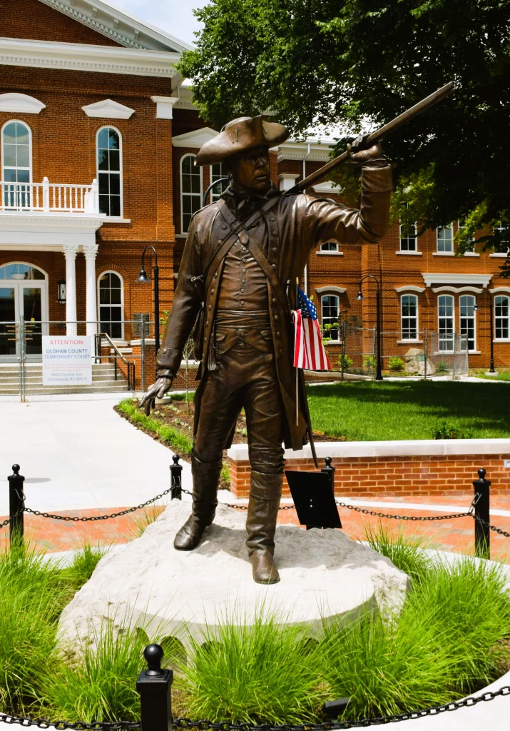 Bronze statue of a Revolutionary War soldier holding a musket in front of a red brick courthouse in La Grange, KY