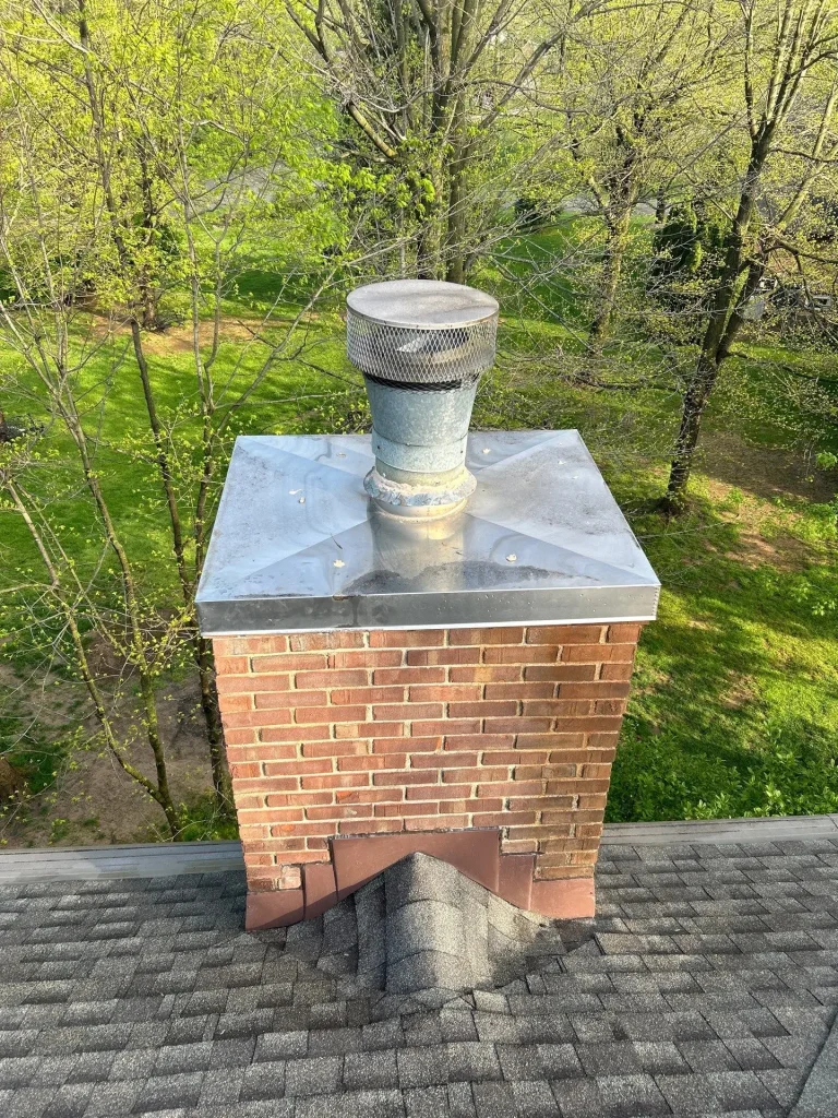 Louisville KY Chimney & Roof Inspection | AIC Roofing & Construction Brick chimney with metal crown and round mesh cap on a gray shingle roof, showing step flashing, counter flashing, and a roof cricket on a Louisville home.