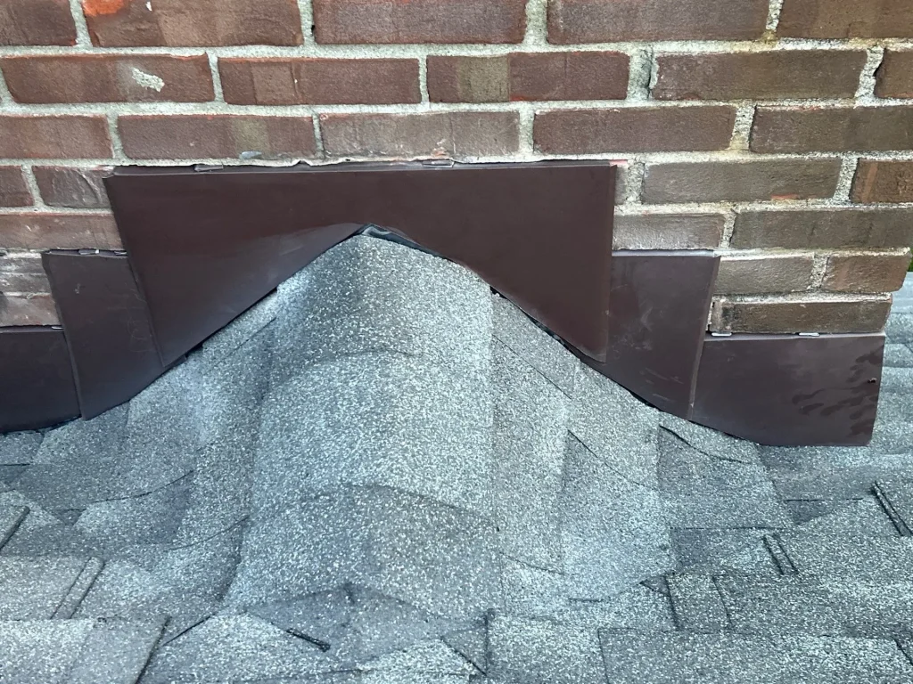 Louisville KY Step Flashing Installation | AIC Roofing & Construction Close-up of a brick chimney base with a shingled roof cricket, brown counter flashing over step flashing on a gray shingle roof in Louisville.