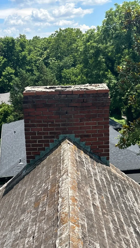 Louisville KY Chimney & Roof Inspection | AIC Roofing & Construction Brick chimney with cracked mortar crown and aging flashing at the roof ridge on a Louisville, KY home, documented by AIC Roofing & Construction.