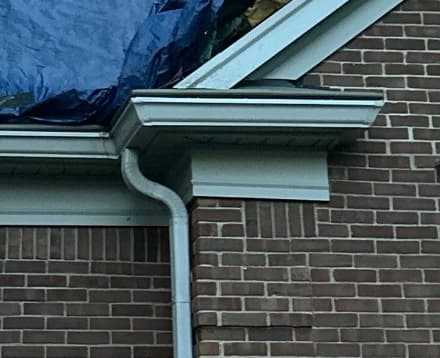 Gutter and downspout at a roof corner on a brick home with a blue tarp covering the roof edge
