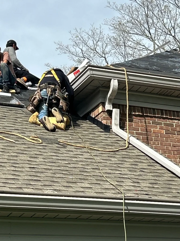Roofers replacing shingles on a Louisville brick home near a roof-to-wall area, with safety harnesses, ropes, and a temporarily removed downspout.