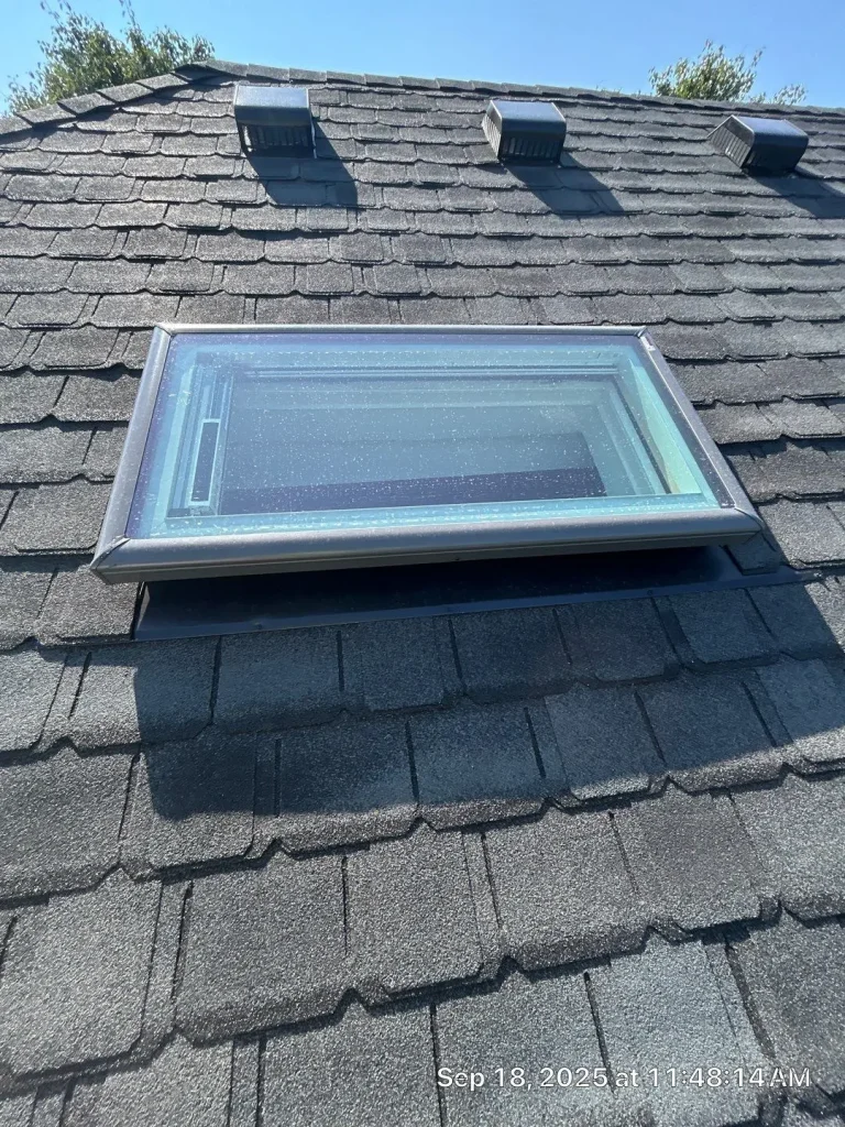 Close-up of a Louisville roof with black shingles, skylight window, and ventilation vents under clear blue sky.