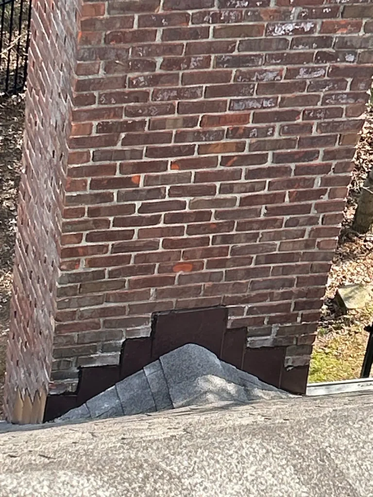 Close-up of a brick chimney with aged step flashing on a shingle roof in Louisville, Kentucky, showing potential water leak areas.