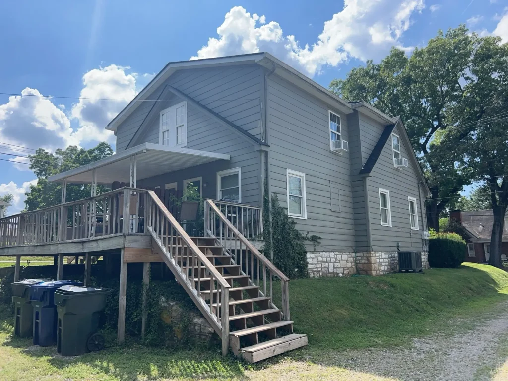 Gray two-story home in Louisville, Kentucky, with horizontal siding, a raised wooden porch, and a stone-and-brick foundation under a bright summer sky.