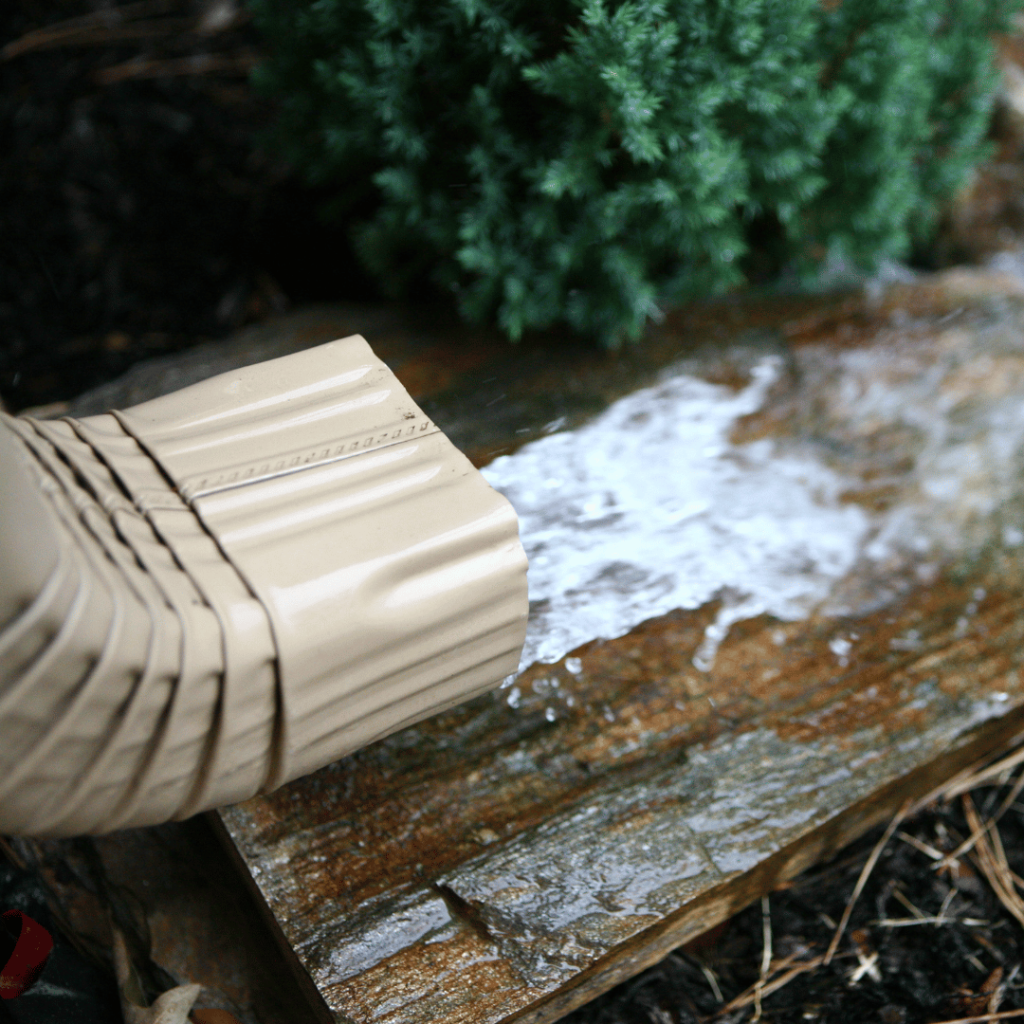 Close-up of a tan aluminum gutter downspout releasing rainwater onto a rock surface near landscaping at a Goshen, Kentucky home.