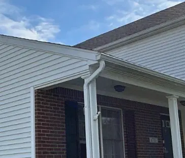 Close-up of white gutters and a downspout attached to the corner of a vinyl-sided Louisville KY home with a brick front porch.