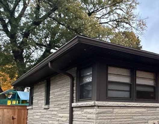 Corner view of a Louisville KY home with newly installed dark gutters, downspout, and soffit above stone exterior walls.
