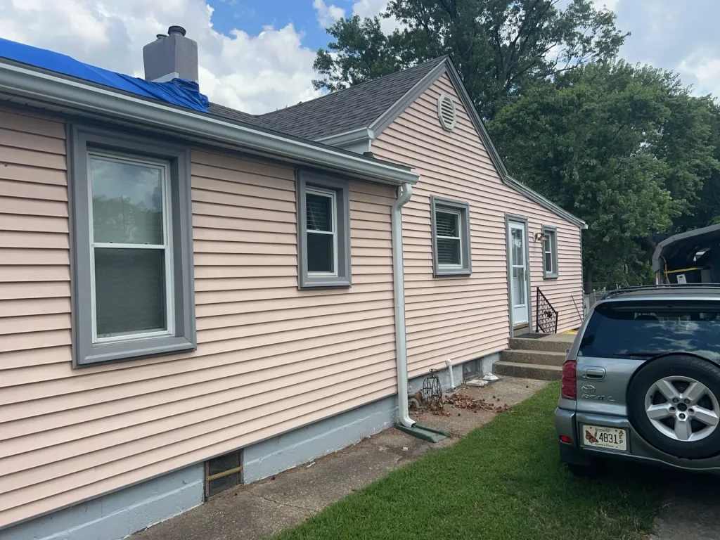 Single-story Louisville KY home with light pink siding and gray trim showing signs of wear, with a blue tarp on the roof and a vehicle parked in the driveway.