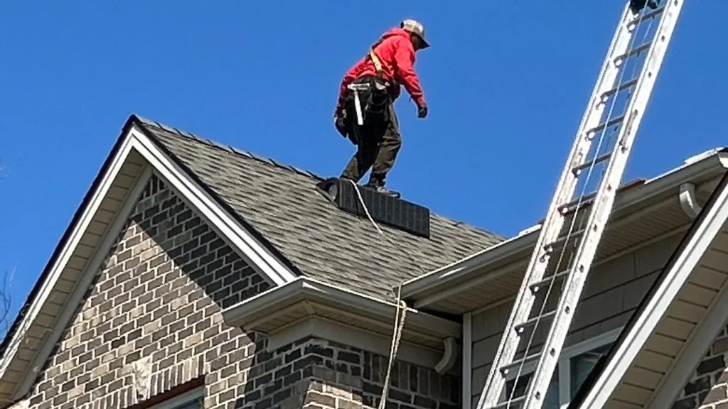 Roofing technician performing asphalt shingle roof repair on residential home in Lexington KY using safety harness near roof peak