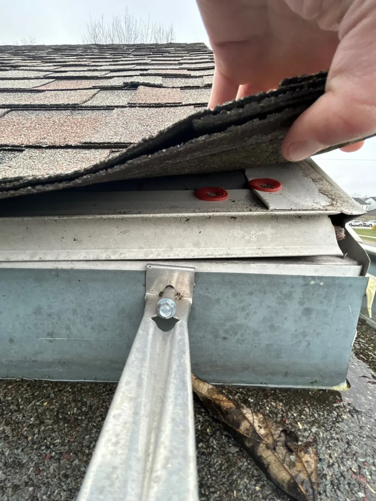 Roof shingles lifted to show gutter apron and drip edge installed above residential gutter system.